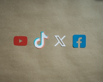 Four social media platform logos are arranged on a brown background. From left to right, the logos are for YouTube in red, TikTok in a combination of red, blue, and white, X in white, and Facebook in blue.